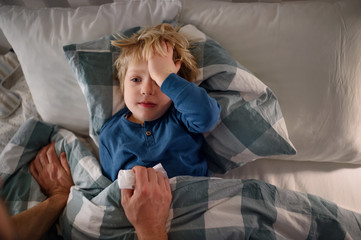 Father checking on small sick son lying in bed indoors at home.
