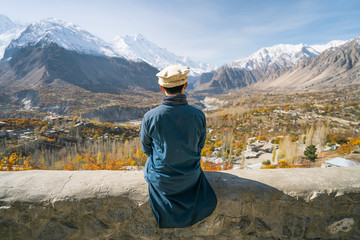 A man with traditional dress sitting on wall and looking at Hunza valley in autumn season, Gilgit Baltistan in Pakistan - obrazy, fototapety, plakaty