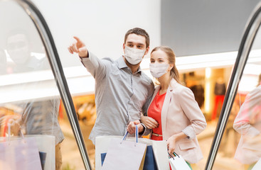 sale, consumerism and pandemic concept - happy young couple wearing face protective medical mask for protection from virus with shopping bags rising on escalator and pointing finger in mall