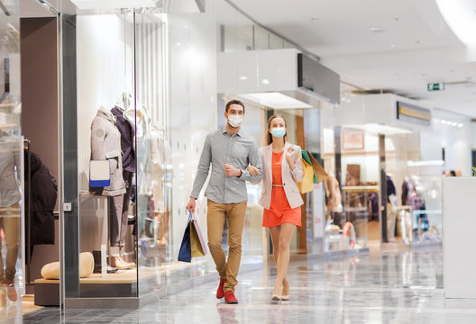 sale, consumerism and pandemic concept - happy young couple wearing face protective medical mask for protection from virus disease with shopping bags walking in mall