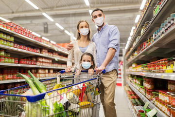 sale, family and pandemic concept - happy mother, father and daughter wearing face protective medical masks for protection from virus disease with shopping cart buying food at supermarket