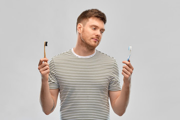 dental care, oral hygiene and people concept - young man comparing wooden and plastic toothbrush over gray background