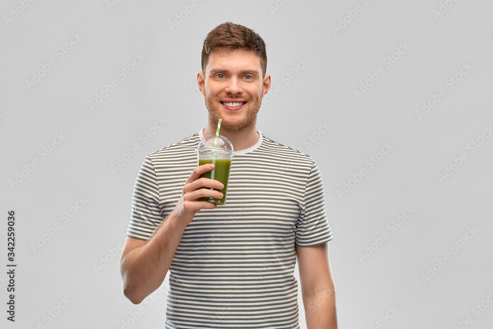 Wall mural healthy eating and people concept - happy smiling man drinking green smoothie from disposable plastic cup with paper straw on grey background