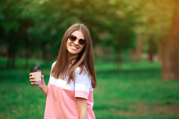 Portrait of beautiful brunette girl in sunglasses walking down the street. Keeping takeaway drink in hand.