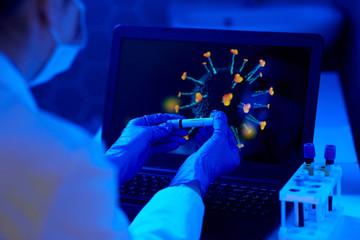 medicine, healthcare and virus concept - close up of hand in protective medical glove holding beaker with coronavirus positive blood test over laptop computer screen at laboratory