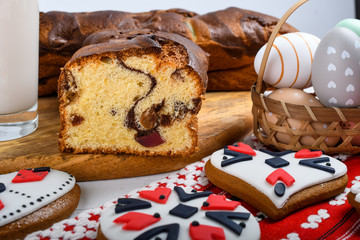 Slices of Romanian sponge cake or chec, cozonac with red traditional towel with manual stitch on wooden board, Easter painted eggs and home made ginger bread hearts, Authentic Easter Brunch concept. 