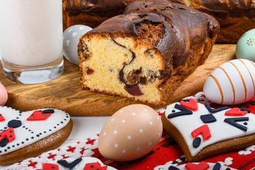 Slices of Romanian sponge cake or chec, cozonac with red traditional towel with manual stitch on wooden board, Easter painted eggs and home made ginger bread hearts, Authentic Easter Brunch concept. 