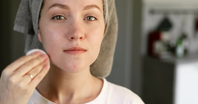 Woman is removing makeup from her face cleaning facial skin, portrait closeup. Girl is wiping face skin looking at camera. Skincare and cleaning procedure at home. Moisturising of skin with lotion.