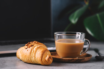 Croissant and cappuccino coffee in glass cup on laptop background.  Home office concept, freelance