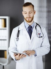 Friendly red-bearded doctor using tablet computer in clinic at his working place. Perfect medical service in hospital and medicine concept