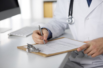 Friendly red-bearded doctor sitting and writing with clipboard in clinic, close-up Medicine concept