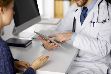 Fototapeta premium Unknown red-bearded doctor and patient woman discussing current health examination while sitting and using tablet computer in clinic, close-up. Medicine concept