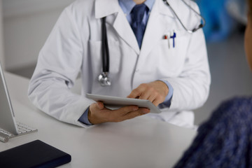 Unknown red-bearded doctor and patient woman discussing current health examination while sitting and using tablet computer in clinic, close-up. Medicine concept