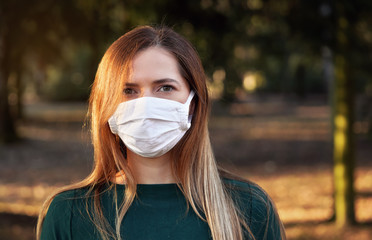 Young woman wearing white cotton virus mouth nose mask, blurred park and trees in background, closeup face portrait
