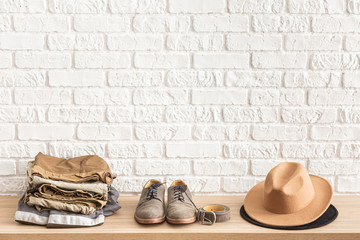 Men's casual outfits on wooden table over white brick wall background with place for text. Shoes, hat, belt and clothes. elegant male accessories. Minimalistic concept of home decor.