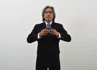 Confident elderly asian businessman in black suit and eyeglasses holding binoculars standing over white wall background, Business vision or marketing research concept