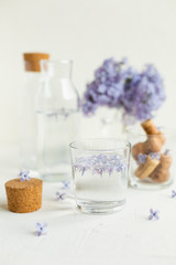 Fototapeta na wymiar A glass of lilac water on the white table. Fresh spring flowers and pure water, healthcare concept.