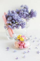Some meringue kisses in a glass jar on the wait background decorated with bouquet of fresh lilac flowers. Spring season.