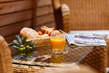 Fresh pastries, orange juice, an olive branch, and a fresh newspaper sit on a wicker table in the courtyard