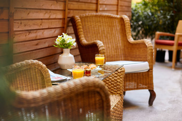 A table in the courtyard is served for Breakfast and decorated with a vase of flowers. On a wicker table there is orange juice, pastries and jam, and a fresh press