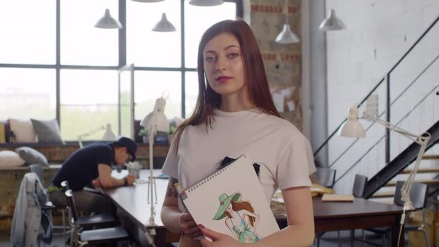 Beautiful female designer holding album with fashion illustration and looking at camera while posing in sewing studio. Suitable for deepfake generation or face swap