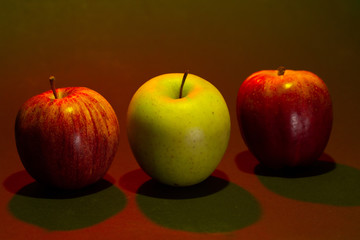 Three apples in a studio photo. Two red ones and a green one. The set light is red except for the green that lights the green apple  from above.