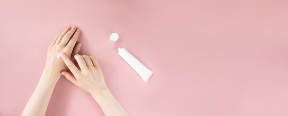 Female hands applying moisturizing cream on skin. Flat lay with white squeeze plastic tube on pink background top view with copy space. Beauty skincare, natural cosmetic. Banner template. Stock photo.