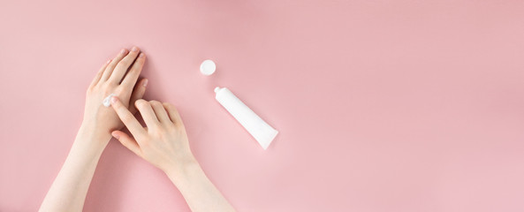 Female hands applying moisturizing cream on skin. Flat lay with white squeeze plastic tube on pink...