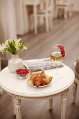 A glass of white wine, crispy croissants, a plate of fresh currant berries and a vase of white flowers are on the table in the cozy living room