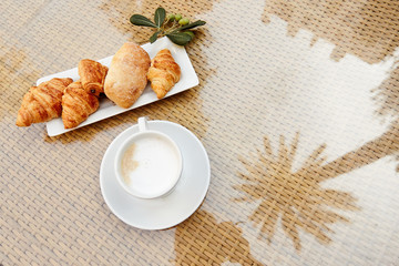 Fresh pastries and a Cup of hot coffee are on the table on the terrace, next to a sprig of olive. View from, space for text