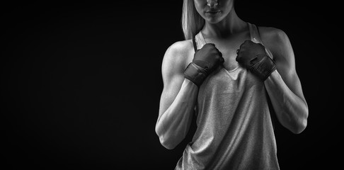 Fototapeta na wymiar Black and white portrait of young woman with muscular body standing against black background. Image of fitness woman in sports clothing and gloves.