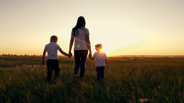 A young woman mother walks with two young children on the grass in the Park. A happy family walks outdoors in the setting sun.
