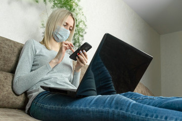 Young blonde woman in a medical mask with a laptop on her lap and a mobile phone in her hands is sitting on the sofa. Concept of work at home, self-isolation, pandemics.