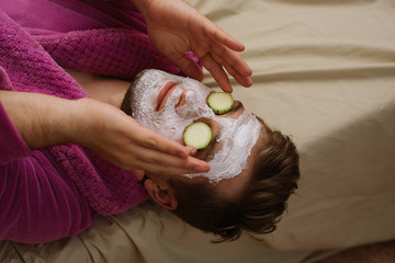 Close up photo of relax man wearing pink robe with facial mask and cucumber on his face laying on...