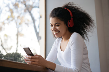 Smiling African American woman using mobile phone, communication, watching videos online. Emotional hipster girl wearing wireless headphones listening to music, sitting at home