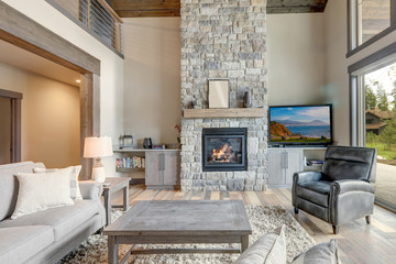 Living room luxury interior with large stone fireplace and leather furniture and huge tall windows with natural tones with TV.