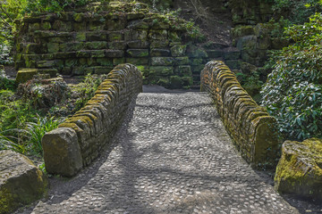 Historical arched stone bridge with cobbled path in Pittencrieff Park, Dunfermline, Scotland with stone wall beyond. Sunny day.