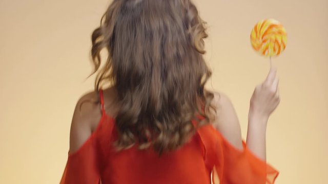 back of cute girl with big caramel candy and curly hair on colored studio background, young woman with sweet lollipop jump up, concept positive mood