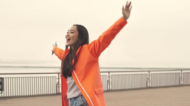 Pretty asian cheery positive woman walking by harbor grant outdoors in orange coat with raised hands.