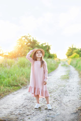 Little girl in dress and straw hat is walking along country road. Happy childhood. Summer vacation. Kid walks along rural road on sunny summer day. Children and nature. Little girl outdoors at sunset