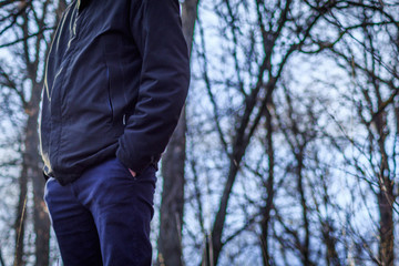 A man in a jacket and jeans stands outdoors in the city park in the evening. His hands in his pockets