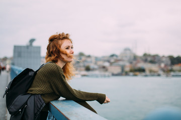 Fototapeta na wymiar Beautiful red-haired curly girl traveler with a backpack on the Galata bridge in Istanbul looks into the distance