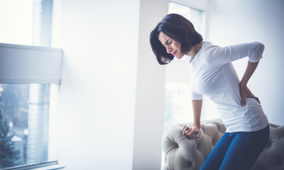 Struggling to walk. Young woman, who has just stood up from sofa, is holding her lower back while...