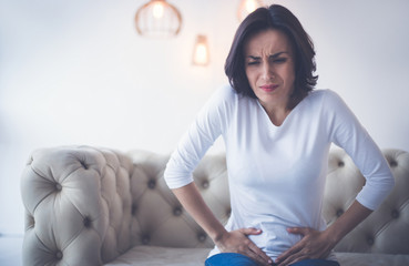 Menstrual pain. Sad woman is sitting on a couch and holding her lower stomach with both hands, while suffering from menstrual cramps.