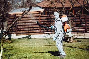 working handyman wearing protective clothing using backpack mistblower for spraying organic pesticides