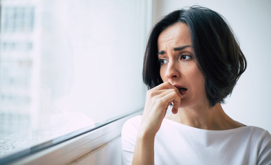 Profound stress. Close-up photo of a sad woman who is looking through the window and biting her nails in distress.