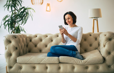 Happy face. Charming young woman is sitting on a sofa in her stylish apartment and smiling, while...