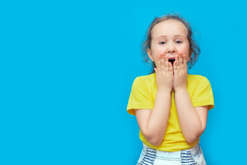 Emotional portrait of a preschool girl on a blue background. A little girl with curly hair opens her mouth wide and screams with delight. He holds his hands to his face. Mock up, copy space