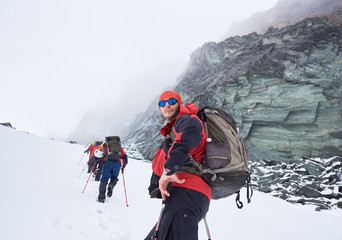 Side view of hiker in sunglasses looking at camera while friends walking on mountain path covered with snow. Male traveler with backpack standing in winter valley with big rock on background.