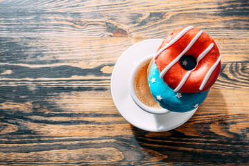 donuts one with blue icing and white stars and with red icing and white stripes and a cup of coffee on a wooden table